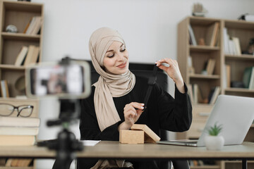 Fototapeta na wymiar Muslim woman doing live stream while unpacking box with new smart watch. Female blogger sharing her feedback about modern gadget with her subscribers.