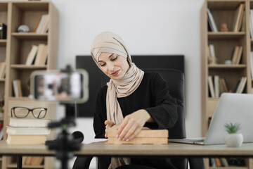 Happy muslim female blogger unpacking gift boxes presents from companies and doing live streaming. Arab woman using smart phone camera for creating content.