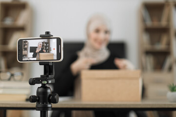 Excited smiling muslim woman in casual wear and hijab recording video on camera while unpacking gift boxes. Female blogger sharing her emotions with her subscribers in social networks.