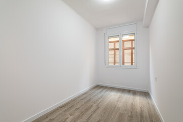 An empty bright room with a window in which the windows of a nearby house are visible. External metal shutters are built into the window to darken the room during daylight hours.
