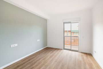 An empty bright room for living with access to balcony and view of neighboring house. Room has transparent glass door with access to balcony. Gray wall and wooden laminate floor create cozy feeling.