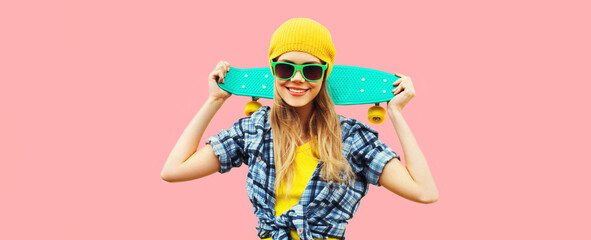 Summer portrait of happy smiling young woman with skateboard wearing colorful clothes on pink background