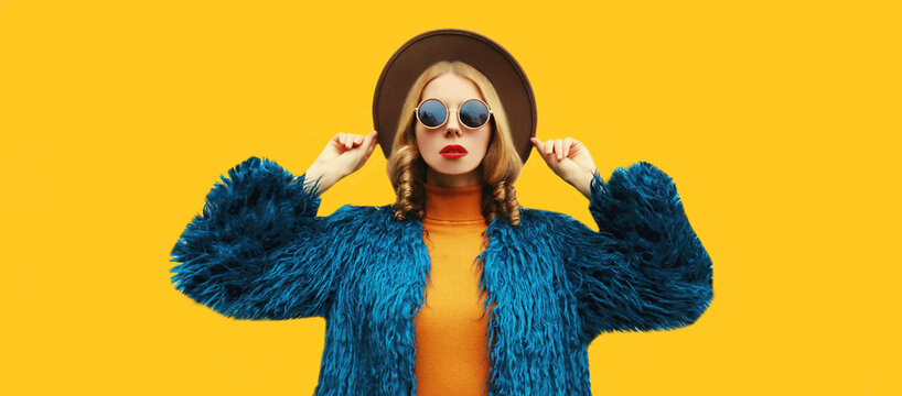 Fashionable portrait of stylish young woman, female model posing wearing blue faux fur, round hat on yellow background