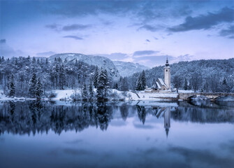 Aerial drone view of beautiful winter landscape with small beautiful church, lake with reflection in water, snowy forest and mountains at sunset top view, Slovenia