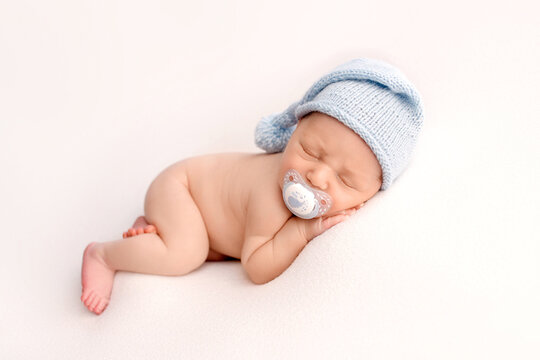 A cute newborn boy in the first days of life sleeps naked with a white pacifier in his mouth on white fabrics in blue knitted woolen cap. Studio professional macro photography, portrait of a newborn.