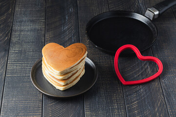 Stack of heart shaped pancakes for romantic breakfast. Valentine's Day concept. Table scene with a сast-iron pan and heart shaped red silicone baking mold. Dark wood background. Love and hearts theme