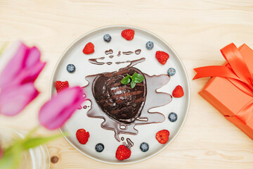 Heart-shaped cake decorated with berries and chocolate icing next to a bouquet of flowers and a gift box. valentine's day gift