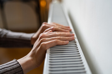 Closeup of woman wants to keep warmth in apartment. Female puts hands on room central heating battery to warm up and prevent illness because of coldness. Person checks work of heater. Selective focus.