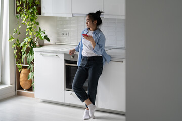 Concerned millennial Asian woman holding mobile phone and looking out window while standing in modern kitchen at home, ordering food online. Sad girl holding smartphone waiting for important call