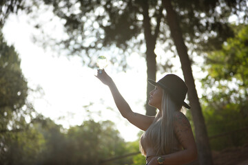Naklejka premium Young, beautiful, blonde woman with hat, holding a light bulb illuminated by the sun passing through it. Concept light, energy, electricity, renewable, solar.