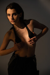 young shirtless woman in black pants and breast tape looking at camera on grey background with lighting.