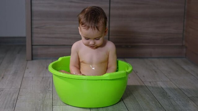 Little cute Caucasian baby sitting in the washbowl and splashing the water. Adorable kid having washing time at home.