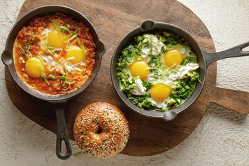 Green and red Shakshuka in cast iron pan with freshly baked bagel on wood cutting board. Poached eggs with tomato sauce or greens. Shakshuka a traditional meal of the Jewish cuisine