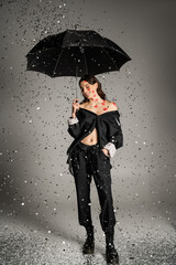happy and stylish woman with red lip prints on body standing with black umbrella under shiny confetti on grey background.