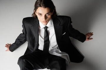high angle view of young stylish woman in black suit and tie looking at camera while sitting on grey background.