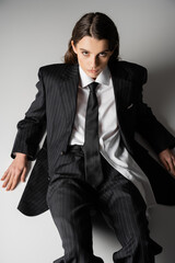 high angle view of fashionable model in oversize shirt and black pantsuit sitting on grey background.