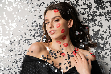 top view of stylish woman with red lip prints lying near festive confetti and looking at camera on grey background.