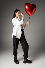 full length of woman in white shirt and black pants with rough boots holding cigarette near red heart-shaped balloon on grey.