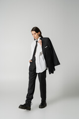 young model in oversize shirt and black pants holding black blazer while walking in rough boots on grey background.