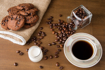 A cup of coffee with chocolate cookie on a dark wooden table. Spilled coffee beans. Milk in a glass jar. Top view.
