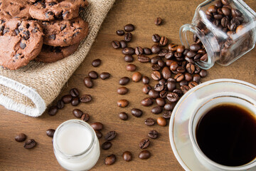 A cup of coffee with chocolate cookie on a dark wooden table. Spilled coffee beans. Milk in a glass jar. Top view.
