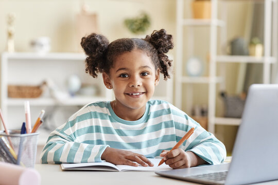 Cute black girl doing homework at desk and smiling at camera in home interior