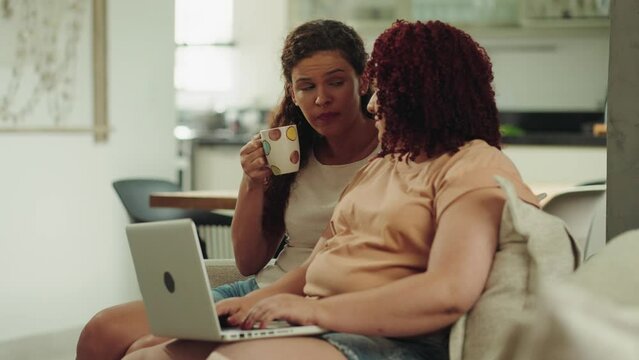 Happy Female Lgbt Couple Relaxing Sitting On Sofa Enjoying Using Laptop Computer, Browsing Internet Choosing Shopping Online Resting Together In Apartment