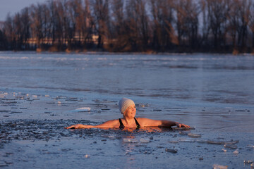 Winter swimming. Mature woman in frozen lake ice hole. Swimmers wellness and endorphin booster swim in cold water. Beautiful female body tempering Cold winter morning landscape. Biohacking routine