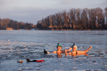 Two women stand at the river's edge as the sun rises, having just finished a winter swim. The cold...