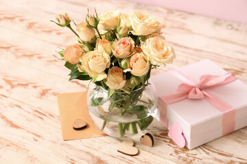 Vase with beautiful rose flowers, envelope and gift box on light wooden background