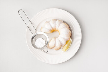 Plate with delicious cake, sieve of powdered sugar and lemon slice on white background