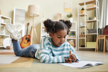 Cute black girl laying on floor at home and writing in notebook or diary