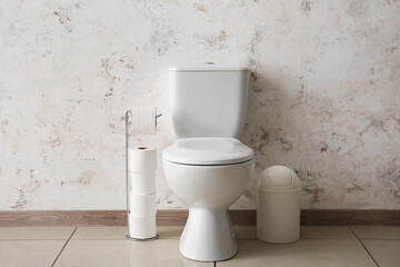 Ceramic toilet bowl, holder with paper rolls and bin near light wall