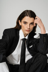 pretty woman in white shirt and black blazer with tie holding hand near head and looking at camera isolated on grey.