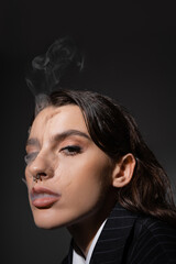 portrait of pretty brunette woman with piercing and makeup smoking and looking at camera isolated on black.