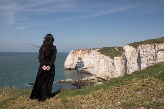 classic rear view portrait of woman in a black dress dress standing on cliffs of sandstone in Etretat, Normandy, France