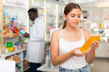 Young woman who came to the pharmacy to buy a hair care remedy chooses a therapeutic shampoo, carefully studying its ..components