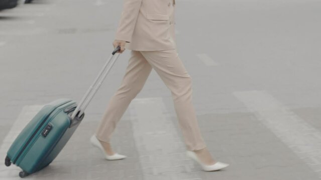 An alluring middle-aged woman in a beige business attire, carrying a green suitcase, walks near a modern airport building