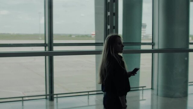 A blonde woman in a blue shirt strolls through an empty airport lounge, with chairs and airport field in the background