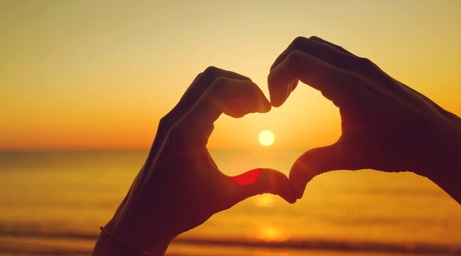 Women's hands in the shape of a heart against the background of the sea and the setting sun let the sun's rays through, toned. Hands in the shape of a heart of love. Romantic background