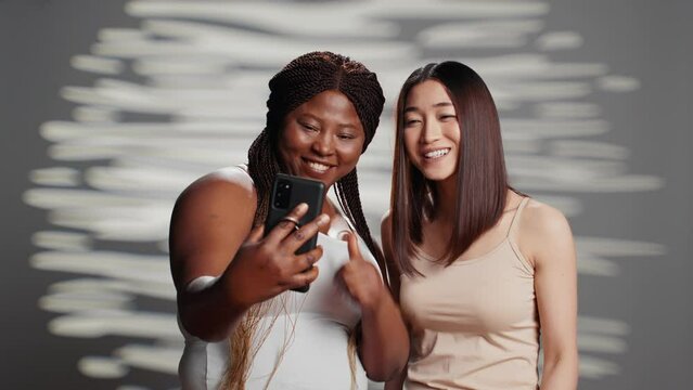 Interracial cheerful ladies taking photos with phone, female models promoting different skintones and body types. Confident natural women laughing with pictures for skincare campaign.