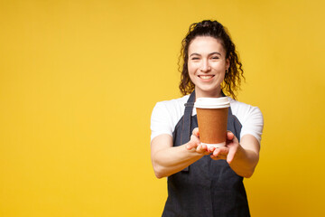 barista girl in a denim apron gives cup of coffee and smiles on yellow background, portrait of...