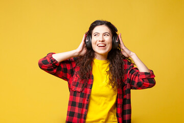 curly brunette girl in a plaid shirt listens to music in headphones on a yellow background and...