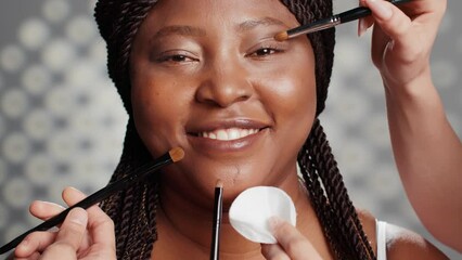 Happy girl receiving makeover from make up artists, team of people using brushes and cotton pads to create natural glowing look on camera. Female model posing for skincare campaign. Close up.