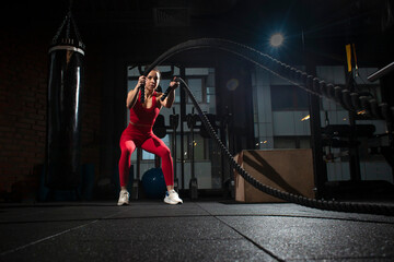 young sports girl in red sportswear trains with ropes in a black dark gym, motivated woman on...