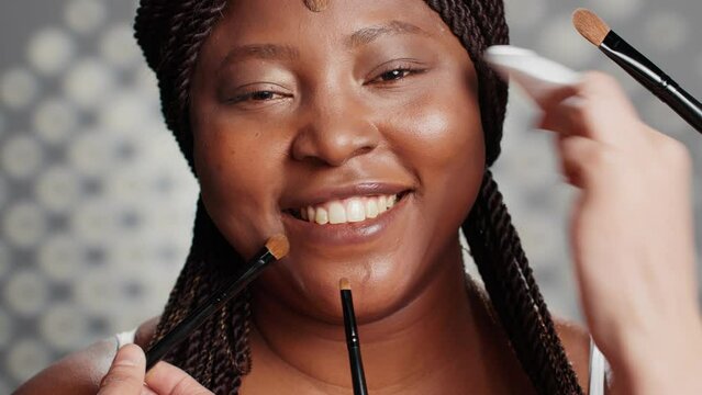 Confident woman having make up done by specialists, team of make up artists using brushes and tools to give model makeover for skincare campaign. People creating luminous radiant look. Close up.