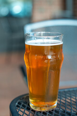 A clear pint drinking glass filled with cold froth from a lager ale. The Irish red ale pint sits on the edge of a metal patio table at a microbrewery. There are tables and chairs in the background.