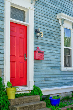 A Colorful Exterior Wall Of A Blue Wooden House With A Single Solid Red Metal Door Has A Small Window Over The Entrance. A Red Metal Post Box Hangs On The Wall. There's A Yellow And Blue Flower Pot.