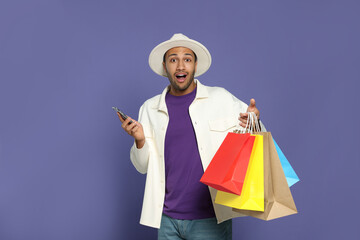 Emotional African American man in hat with shopping bags and smartphone on purple background