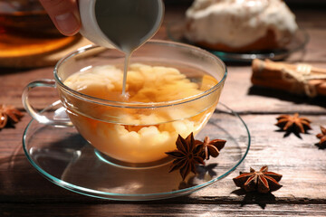 Woman pouring milk into glass cup of tea with anise stars at wooden table, closeup